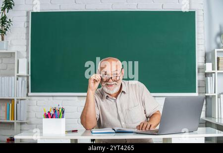 Vecchio insegnante di sesso maschile di fronte alla lavagna. Uomo insegnante seduto di fronte alla tavola verde. Uomo sorridente ritratto in occhiali seduto alla scrivania, in classe. Maschio Foto Stock