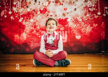 Il sorridente ragazzo con il papillon rosso è decorato a gambe incrociate con decorazioni a cuore Foto Stock