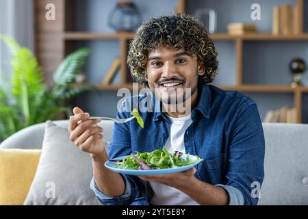 Un uomo allegro siede comodamente su un divano, gustando un'insalata verde fresca. La sua espressione di felicità evidenzia uno stile di vita sano. L'accogliente ambiente del soggiorno aggiunge calore e relax. Foto Stock