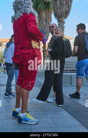 Sabona. Italia - 06 dicembre 2024: Un individuo vestito con un costume teatrale di velluto rosso con scarpe da ginnastica moderne in una piazza pubblica, circondata da c Foto Stock