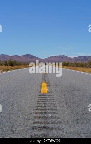 Una lunga e dritta strada asfaltata con linee gialle si estende verso montagne lontane e basse sotto un cielo azzurro nel deserto. Foto Stock