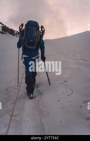 Un alpinista cammina in una squadra di corda con le racchette da neve sullo zaino sul ghiacciaio di Alphubel nel Canton Vallese in Svizzera. Le nuvole grigie sono visibili all'orizzonte. La luce mattutina rossastra è visibile all'orizzonte sopra l'alpinista. Foto Stock