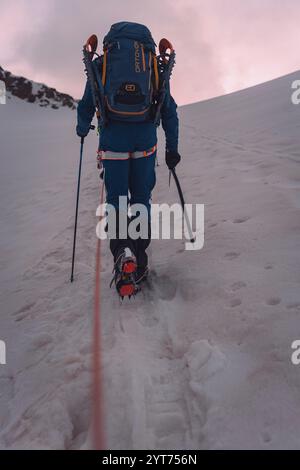 Un alpinista cammina in una squadra di corda con le racchette da neve sullo zaino sul ghiacciaio fino ad Alphubel, in Svizzera, nel Canton Vallese. Le nuvole grigie sono visibili all'orizzonte. La luce mattutina rossastra è visibile all'orizzonte sopra l'alpinista. Foto Stock