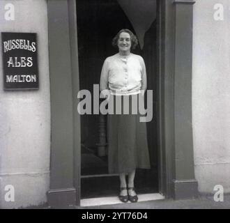 Anni '1950, storica, una signora che si ferma per una foto all'ingresso di un pub, Star Inn, rivenditore autorizzato di birre, vini, liquori e tabacco, una Samuel Cherry. Il cartello sul muro dice Russells Ales Malton, North Yorkshire, Inghilterra, Regno Unito, che risale al 1771. Russells & Wrangham Ltd, Derwent Brewery, Castlegate, Malton, North Yorkshire nacque nel 1897 con la fusione di James Russell & Son e William Wrangham Ltd Foto Stock