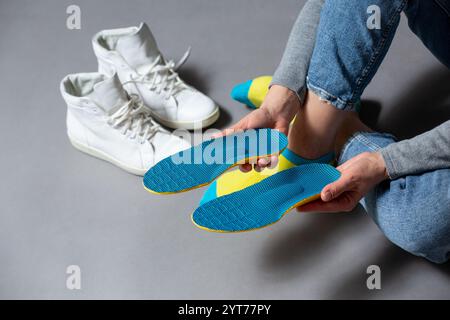 Primo piano di mani femminili con solette ortopediche blu, sneakers bianche su sfondo grigio . Assistenza sanitaria e trattamento ortopedico e prevenzione di Foto Stock