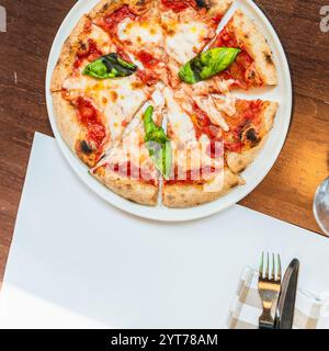 Pizza margherita a fette con mozzarella fusa e foglie di basilico su un piatto bianco, servita su un tavolo di legno del ristorante Foto Stock
