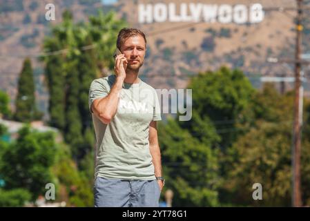 Un uomo si trova all'aperto a Los Angeles, California, a parlare al cellulare. L'iconica insegna di Hollywood è visibile sullo sfondo, circondata da lussureggianti giardini Foto Stock