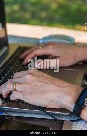 Primo piano delle mani di una donna anziana che digita sulla tastiera del notebook, lavorando in remoto in ambienti esterni Foto Stock
