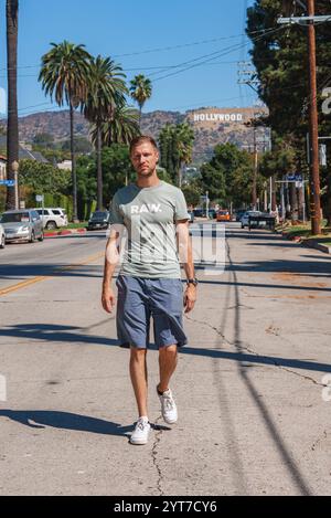 Un uomo con una maglietta verde chiaro cammina lungo una strada costeggiata da palme a Los Angeles. L'iconica insegna di Hollywood è visibile contro un cielo azzurro. Foto Stock