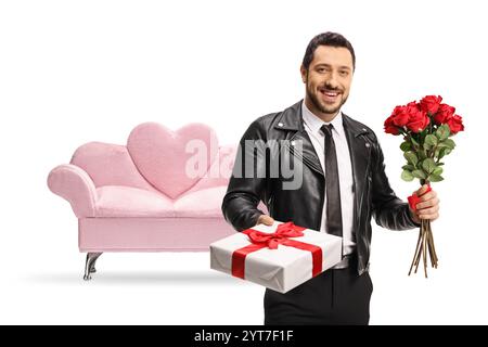 Uomo in una giacca di pelle con una confezione regalo e un mazzo di rose rosse davanti a un divano in velluto rosa isolato su sfondo bianco Foto Stock