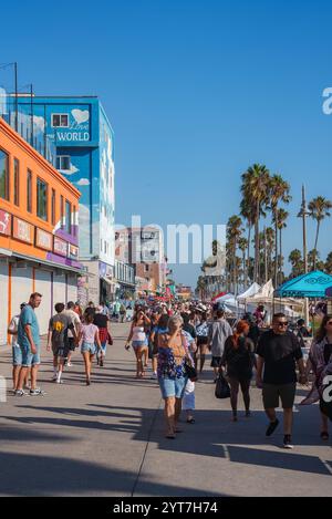 La passerella di Venice Beach è piena di gente, negozi colorati e palme. Un murale recita "Love World" sotto un cielo azzurro. Foto Stock