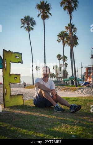 Un uomo siede in un'area erbosa a Venice Beach, Los Angeles, con alte palme e una grande scultura che ricorda una lettera e sullo sfondo. Foto Stock