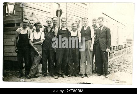 Cartolina originale degli anni '1920 di uomini della classe operaia - costruttori o navvii, costruzione di capanne di legno, attrezzi, forse il caposquadra che indossa un vestito e una cravatta alla fine della fila. Alcuni uomini che indossano berretti piatti, alla fine degli anni '1920 o all'inizio degli anni '1930 nel Regno Unito Foto Stock