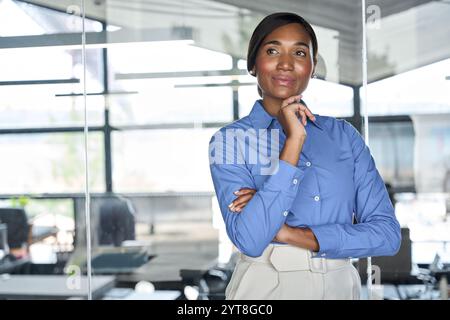 Giovane donna d'affari afro-americana in piedi in ufficio che guarda lontano pensando. Foto Stock