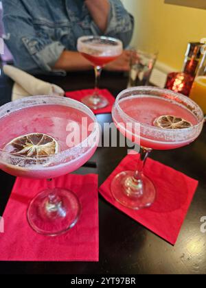 Due eleganti bicchieri di cocktail rosa guarniti con fette di agrumi essiccate su tovaglioli rosa brillanti, con una persona seduta sullo sfondo. Foto Stock