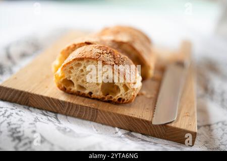 Fette di pane croccante ciabatta con semi di sesamo su un asse di legno con coltello. Primo piano con profondità di campo ridotta. Foto Stock