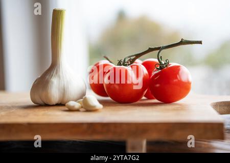 Pomodori rossi maturi e aglio fresco su un tagliere di legno di fronte a una luminosa finestra della cucina. Sfondo con breve profondità di campo e spazio per il testo. Primo piano Foto Stock