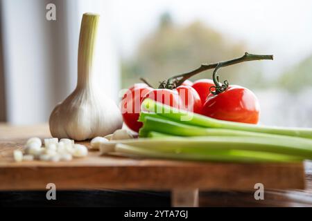 Pomodori rossi con aglio fresco e cipolle primaverili su un tagliere di legno davanti alla finestra della cucina. Profondità di campo ridotta. Foto Stock