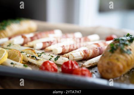 Asparagi bianchi fritti avvolti in pancetta con erbe, patate e pomodori su una teglia da forno. Profondità di campo ridotta. Foto Stock