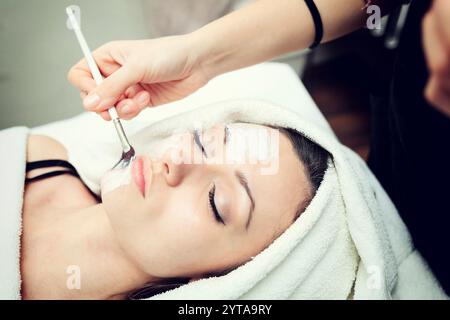 Cosmetico applicando bellezza viso maschera per il giovane bella donna a spa salone Foto Stock