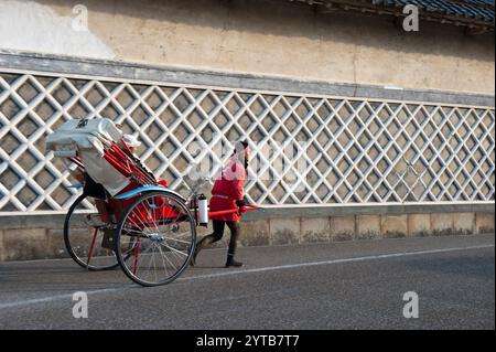 Una jinrikisha di pedicab a propulsione umana con un cliente a bordo supera un tradizionale schema murale a Hikone, Shiga, Giappone. Foto Stock