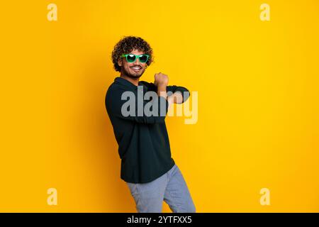 Giovane uomo in camicia nera e jeans che posa giocosamente con occhiali da sole verdi su sfondo giallo brillante Foto Stock