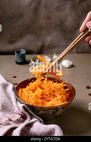 Cucina coreana e asiatica. La mano dell'uomo utilizza le bacchette per raccogliere l'insalata di verdure di carote marinate con l'aglio dal recipiente in ceramica. Condimento piccolo Foto Stock