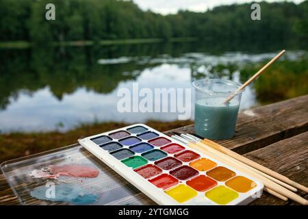 Primo piano dell'installazione dell'acquerello con i pennelli e il vaso d'acqua aquarelle poggiati su un tavolo di legno vicino a un lago forestale - concetto di scincisione ad aria compressa Foto Stock