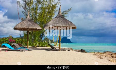 Spiaggia tropicale con sedie a sdraio e ombrelloni di paglia sulle nuvole di tuono e penisola le Morne Brabant sullo sfondo. Inverno sull'isola di Mauritius Foto Stock