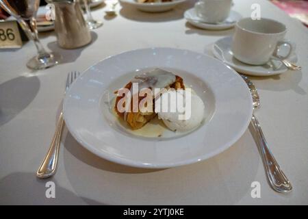 Un piatto di strudel di mele condito con salsa alla vaniglia, servito insieme a una paletta di gelato alla vaniglia su un tavolo bianco. Foto Stock