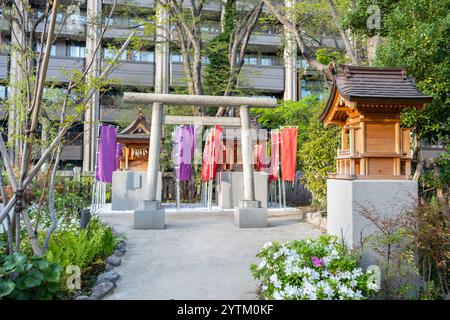 Tradizionale santuario shintoista con porta torii in pietra e striscioni rosso e viola, sfondo di edifici moderni della città di Tokyo. Giappone Foto Stock