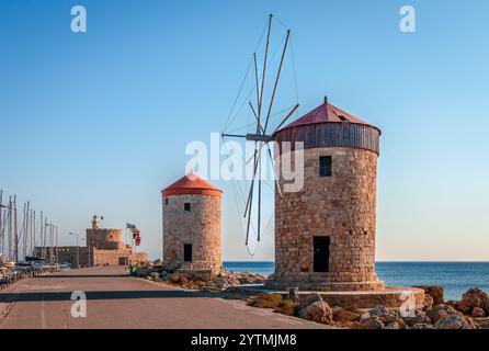 I mulini a vento medievali sul ponte delle onde al porto di Mandraki, Rodi, Grecia. La fortezza e il faro di Agios Nikolaos si trovano nel backgorund. Foto Stock