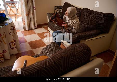 Una donna siede su un divano, giocando un sitar in un soggiorno decorato con calore. L'atmosfera è tranquilla e rilassata, con luce naturale che scorre in fro Foto Stock