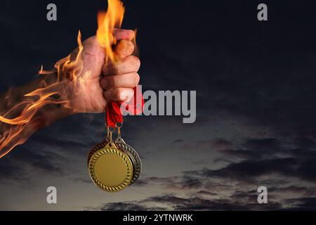 Uomo con medaglie d'oro, d'argento e di bronzo nella sua mano fiammeggiante contro il cielo oscuro, primo piano Foto Stock