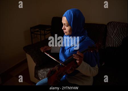 Una donna che indossa un hijab blu siede in una stanza poco illuminata tenendo in mano un violino, creando un'atmosfera serena e introspettiva. Foto Stock