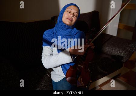 Una donna che indossa un hijab blu tiene un violino, godendo di un momento di pace. I suoi occhi sono chiusi mentre si siede su un comodo divano, trasuda tranquillità e Foto Stock