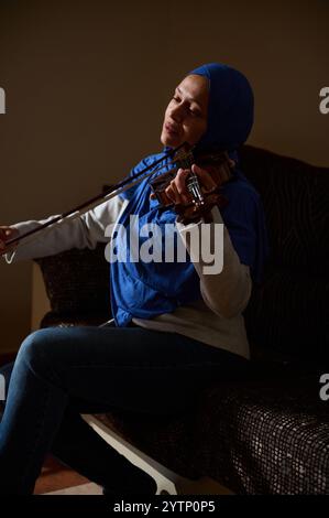 Una donna che indossa un hijab blu siede su un divano, suona con passione il violino, immersa nella musica, catturando un momento di profonda concentrazione ed emozione Foto Stock