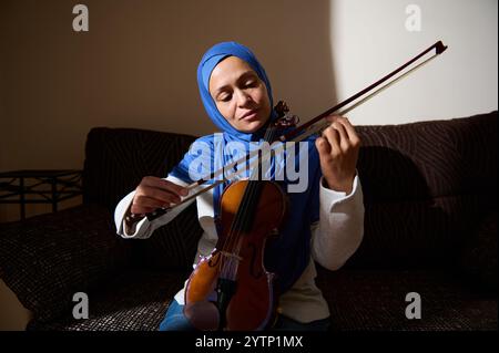 Una donna che indossa un hijab blu si siede su un divano, suona il violino con concentrazione ed emozione in una stanza calda illuminata. Foto Stock