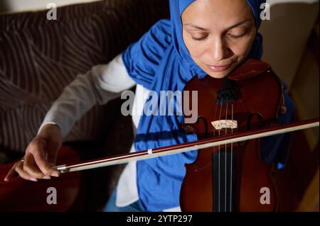 Una donna che indossa un hijab blu che suona abilmente il violino, mettendo in risalto il suo talento musicale e concentrandosi in un ambiente interno con illuminazione naturale. Foto Stock