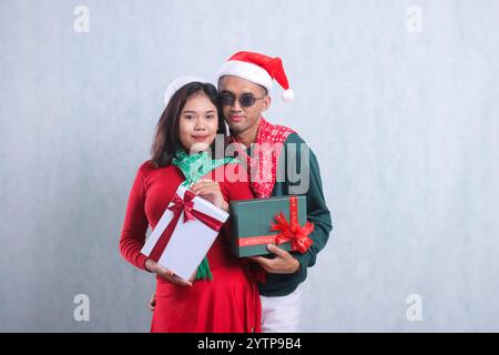 ritratto di una giovane coppia che indossa maglioni natalizi, cappelli e sciarpe di babbo natale, sorridendo elegantemente guardando avanti portando con sé un regalo di natale rosso e bianco Foto Stock