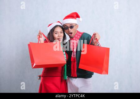 ritratto di una giovane coppia che indossa maglioni natalizi, cappelli di babbo natale e sciarpe, sorpreso guardando la macchina fotografica entrambe in possesso di un sacchetto di carta rosso con regali di natale Foto Stock