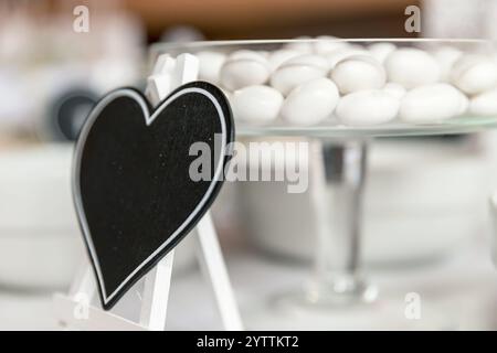 Piatto di vetro pieno di mandorle bianche zuccherate, esposto su un tavolo a buffet durante un ricevimento di nozze. Piccola tavola vuota a forma di cuore per il testo. Foto Stock