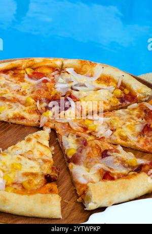 Pizza tagliata a fette sullo sfondo di una piscina blu. Deliziosa pizza con prosciutto, mais, cipolle, pomodori e formaggio. Foto Stock