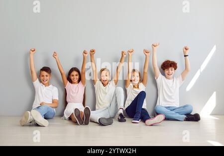 Bambini felici seduti sul pavimento su sfondo grigio con le mani in alto e guardando la macchina fotografica. Foto Stock