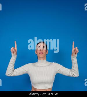 Giovane donna che punta verso l'alto con entrambe le dita indice che guardano verso l'alto su uno sfondo blu Foto Stock