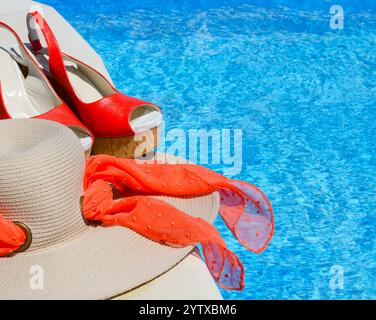 Cappello e sandali da donna accanto alla piscina. Bellissimi sandali rossi e paglia su uno sfondo di acqua blu Foto Stock