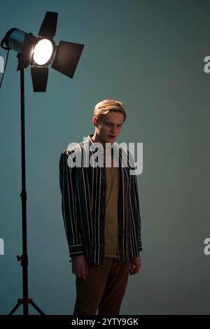 Giovane uomo biondo in camicia a righe in piedi in studio in luce soffusa Foto Stock