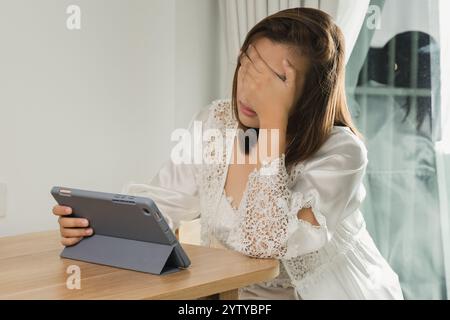 Una donna asiatica in un abito da notte in raso bianco prova dolore agli occhi e fatica mentre legge il suo programma di lavoro su un tablet, facendola stare al passo con lei Foto Stock