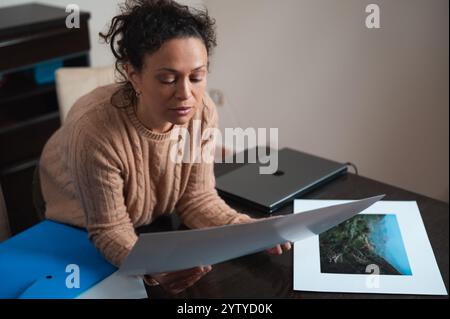 Una donna con un maglione sta esaminando attentamente le vivaci fotografie di paesaggi a una scrivania. Foto Stock