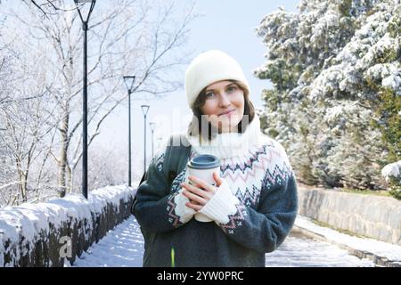 Una donna vestita con un maglione invernale tiene in mano una tazza con una bevanda calda. Donna sullo sfondo del parco invernale. Foto Stock